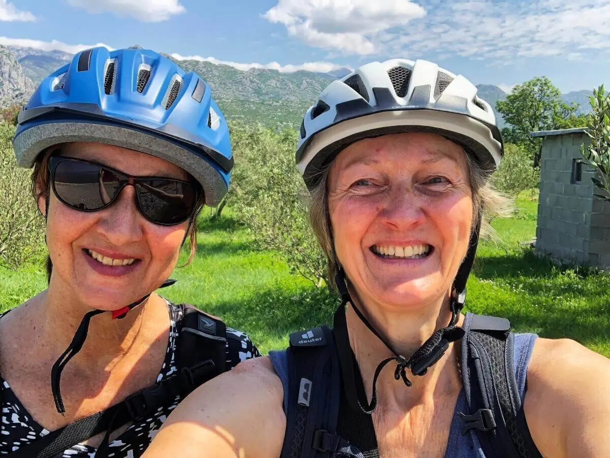 Zwei Personen mit Fahrradhelmen und Rucksäcken stehen auf einer Wiese vor Bergen und bewölktem Himmel. | © DAV Markt Schwaben | Foto Hildegard Petschik
