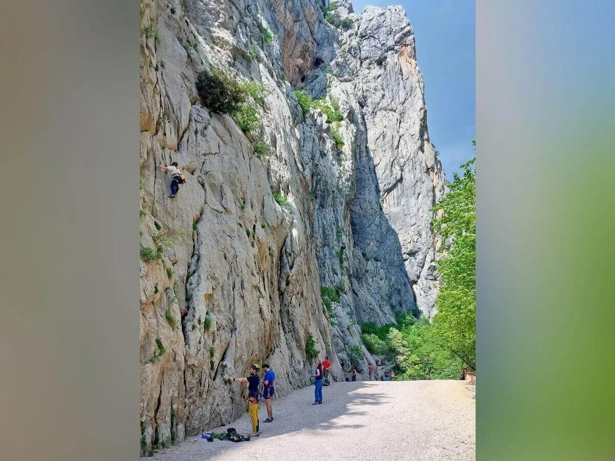 Eine Person klettert an einer hohen Felswand, andere stehen unten bereit; schroffe Felsen und Vegetation umgeben die Szene. | © DAV Markt Schwaben | Foto Lutz Gründel