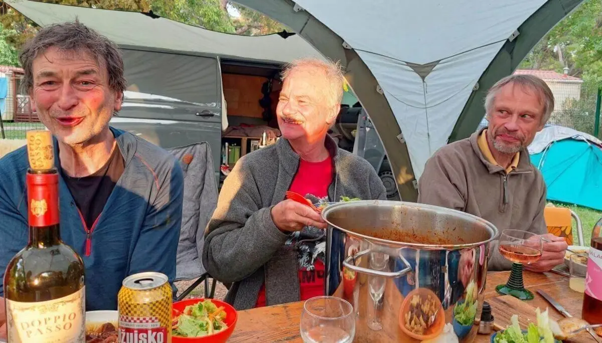 Drei Personen sitzen vor Zelt und Camper am gedeckten Tisch mit Wein, Bier und Eintopf; entspannte Atmosphäre. | © DAV Markt Schwaben | Foto Lutz Gründel