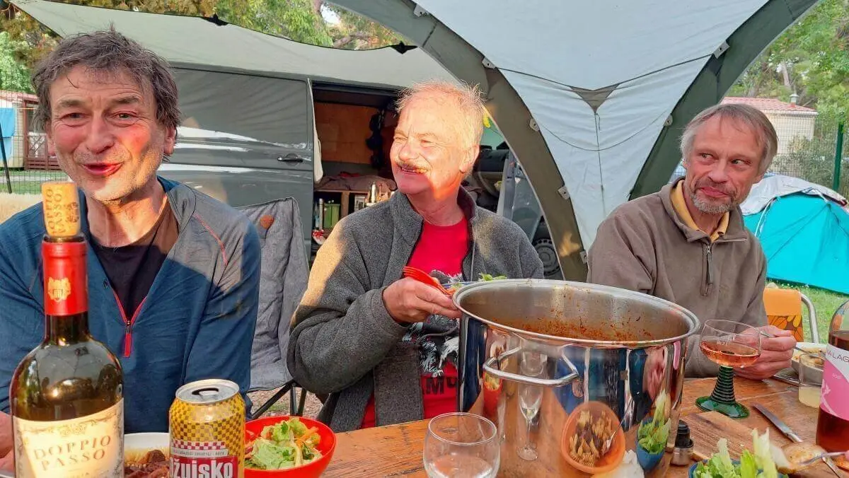 Drei Personen sitzen vor Zelt und Camper am gedeckten Tisch mit Wein, Bier und Eintopf; entspannte Atmosphäre. | © DAV Markt Schwaben | Foto Lutz Gründel