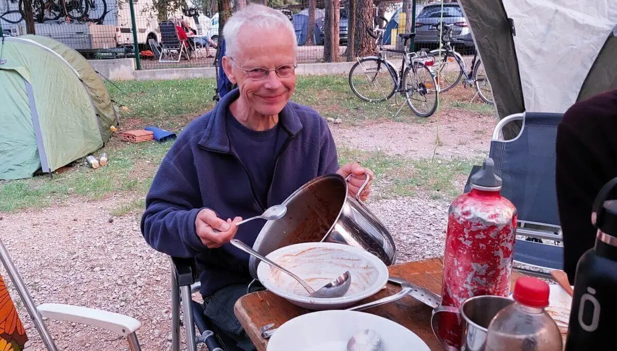 Eine Person hält einen leeren Kochtopf und eine Kelle am Campingtisch; Zelte und Fahrräder im Hintergrund. | © DAV Markt Schwaben | Foto Lutz Gründel