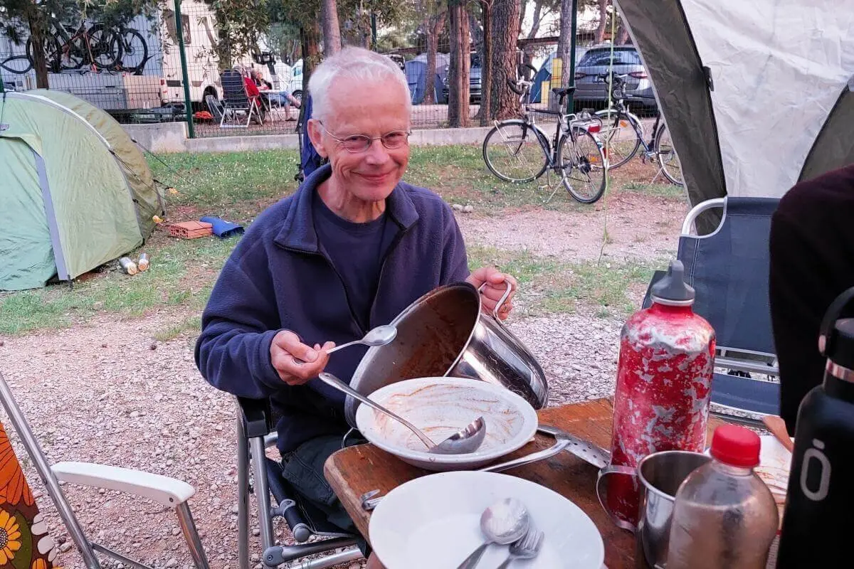 Eine Person hält einen leeren Kochtopf und eine Kelle am Campingtisch; Zelte und Fahrräder im Hintergrund. | © DAV Markt Schwaben | Foto Lutz Gründel