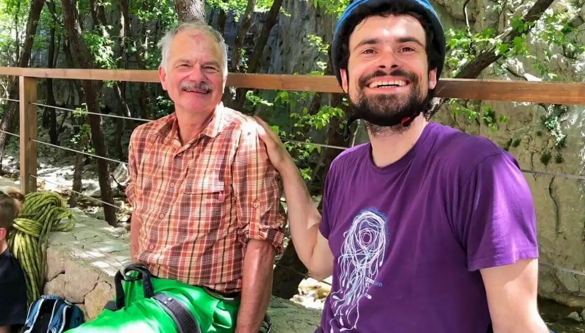 Zwei Personen sitzen entspannt im Wald mit Kletterausrüstung; Felsen und Holzgeländer im Hintergrund. | © DAV Markt Schwaben | Foto Hildegard Petschik