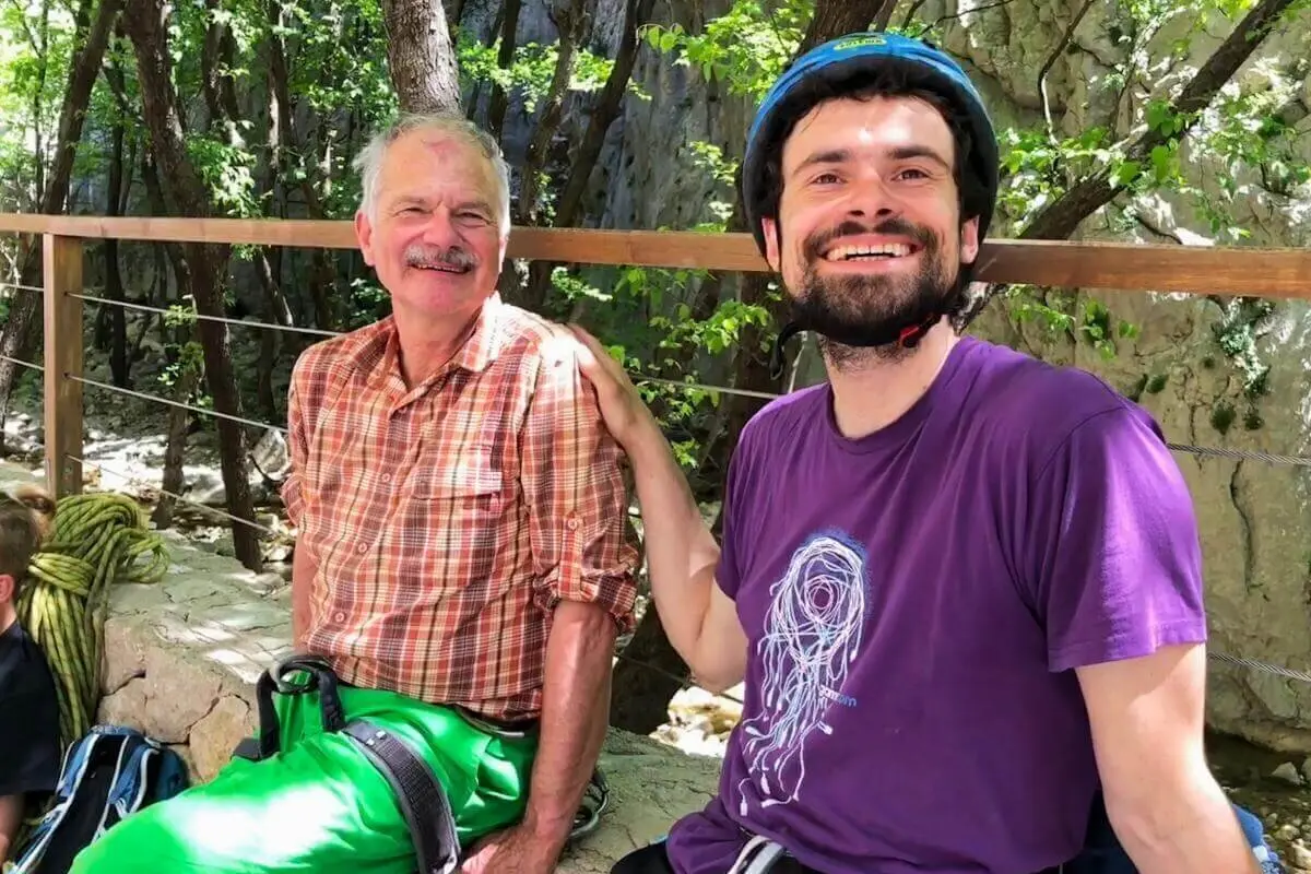Zwei Personen sitzen entspannt im Wald mit Kletterausrüstung; Felsen und Holzgeländer im Hintergrund. | © DAV Markt Schwaben | Foto Hildegard Petschik