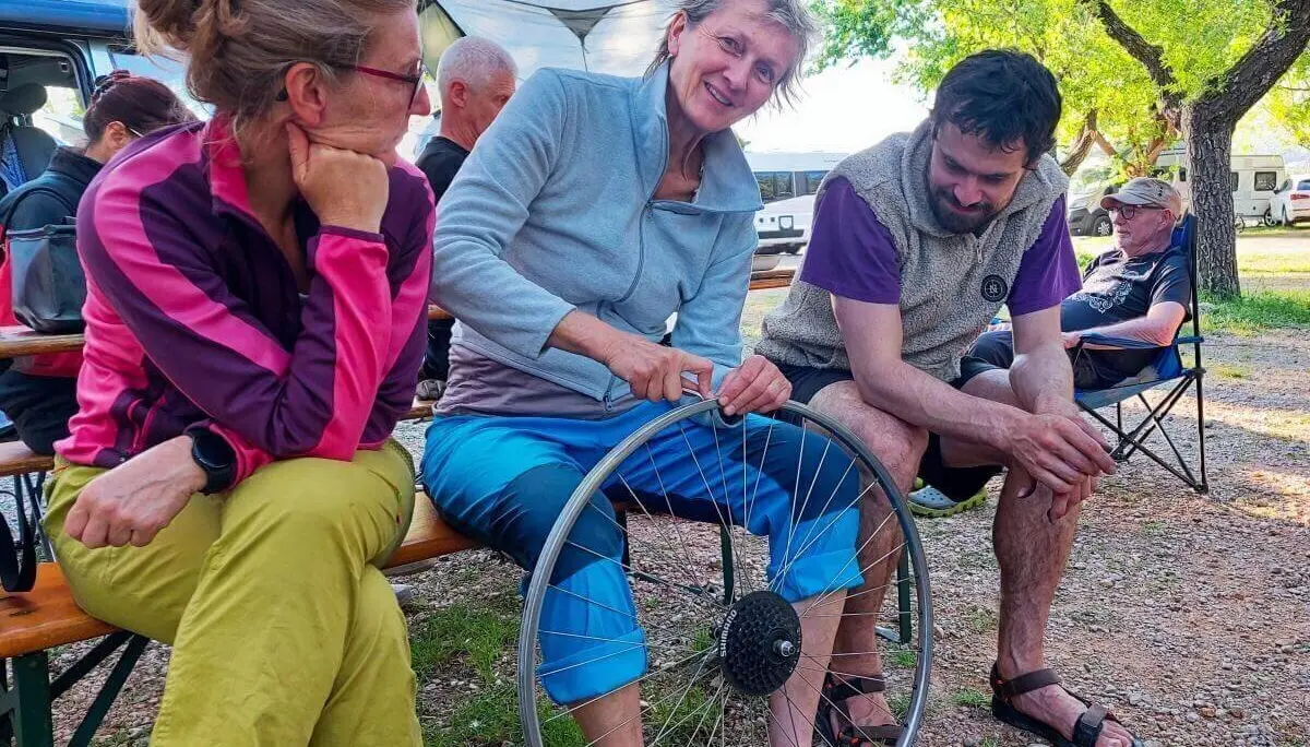 Eine Person zeigt ein Fahrradteil in einer Gruppe; Campervans und Bäume rahmen die Szene. | © DAV Markt Schwaben | Foto Lutz Gründel