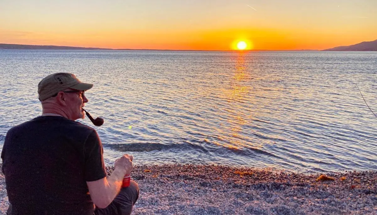 Eine Person sitzt rauchend am Wasser bei Sonnenuntergang; Felsen, Dosen und goldene Lichtstimmung. | © DAV Markt Schwaben | Foto Stefan Schütz