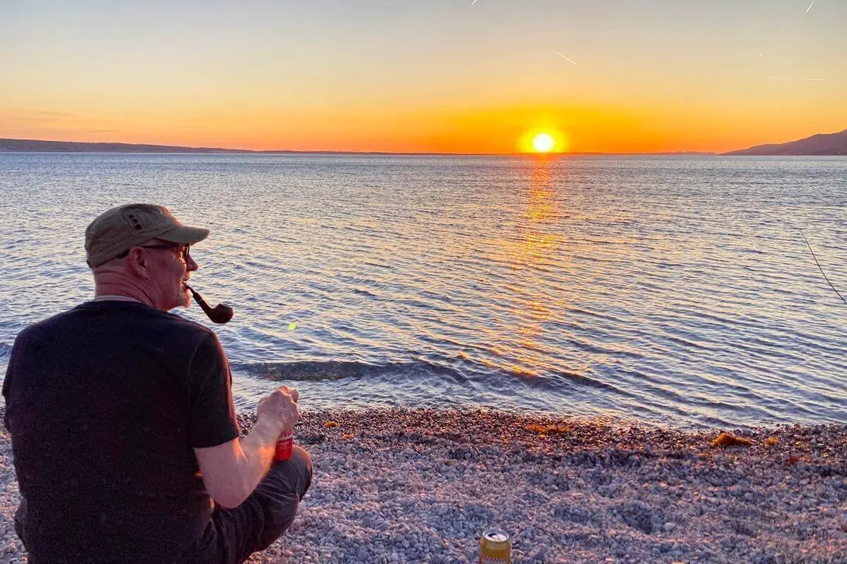 Eine Person sitzt rauchend am Wasser bei Sonnenuntergang; Felsen, Dosen und goldene Lichtstimmung. | © DAV Markt Schwaben | Foto Stefan Schütz