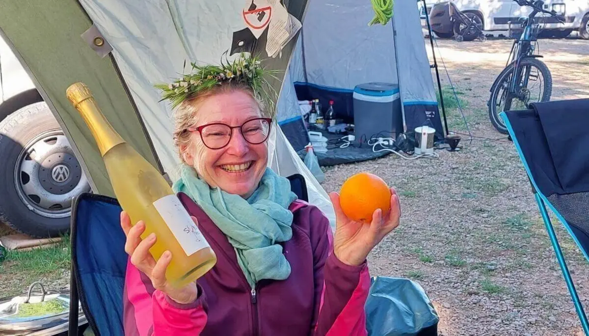 Eine Person mit Blätterkrone hält Sekt und Orange; Zelte, Fahrräder und Fahrzeuge im Hintergrund. | © DAV Markt Schwaben | Foto Lutz Gründel