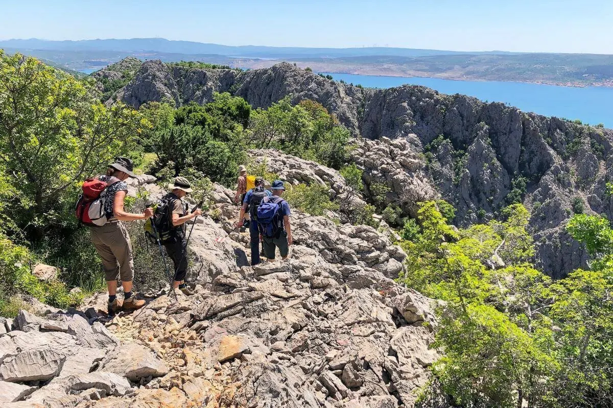 Fünf Wandernde auf felsigem Pfad mit Blick auf das Meer und entfernte Berge unter blauem Himmel. | © DAV Markt Schwaben | Foto Hildegard Petschik