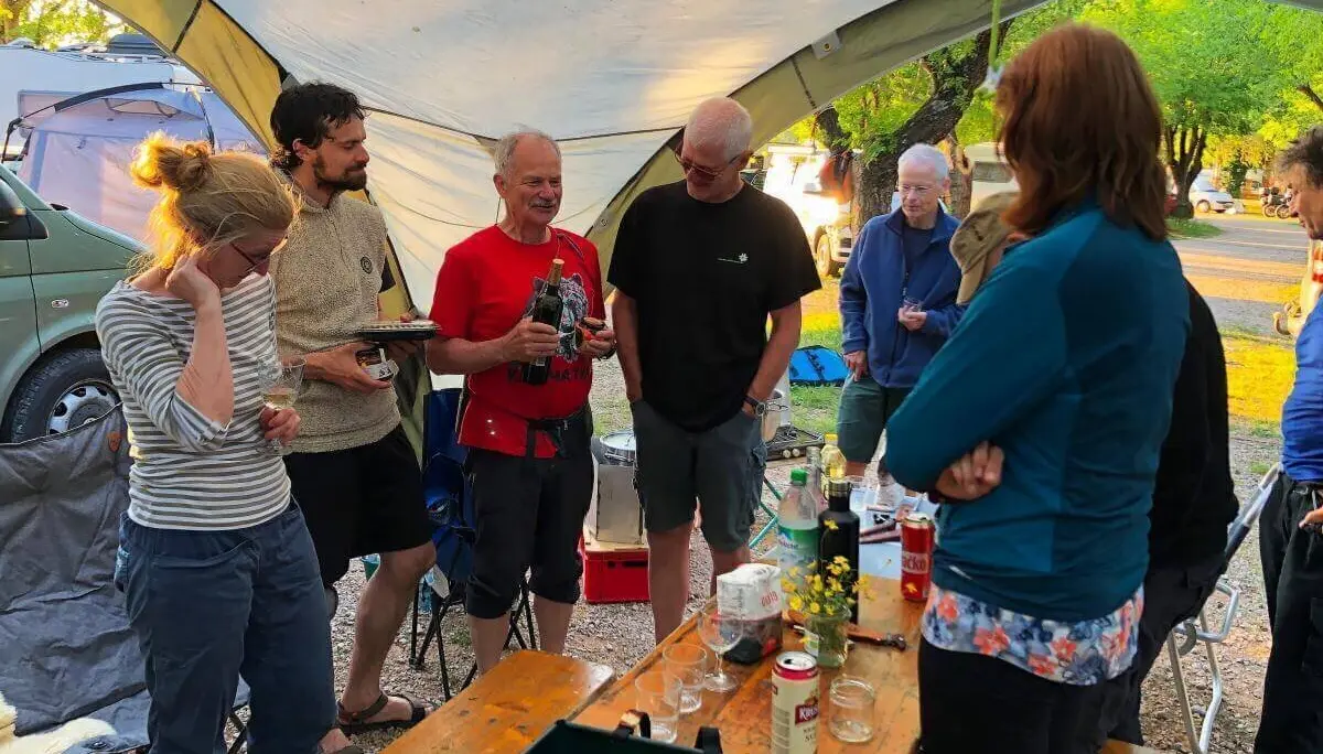 Acht Personen stehen oder sitzen am Tisch mit Getränken und Pflanze; Zelte und Van im Hintergrund. | © DAV Markt Schwaben | Foto Hildegard Petschik