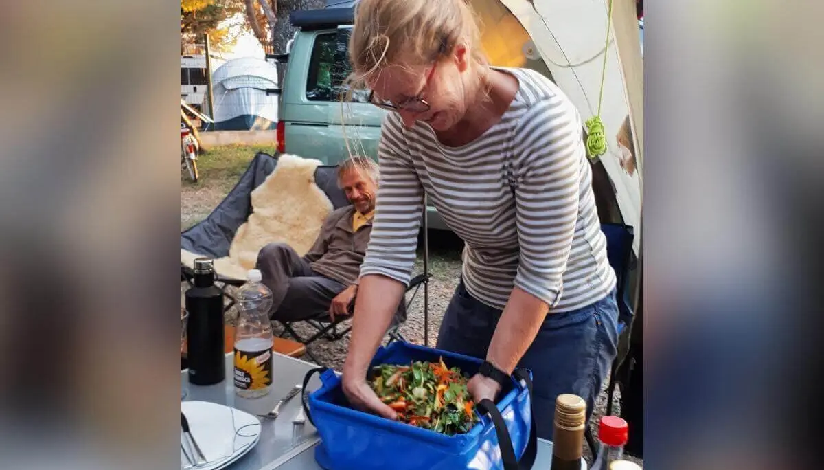 Eine Frau mischt Salat am Campingtisch; Geschirr, Weinflaschen und Zelte rahmen die Szene. | © DAV Markt Schwaben | Foto Martin und Silvia Mack