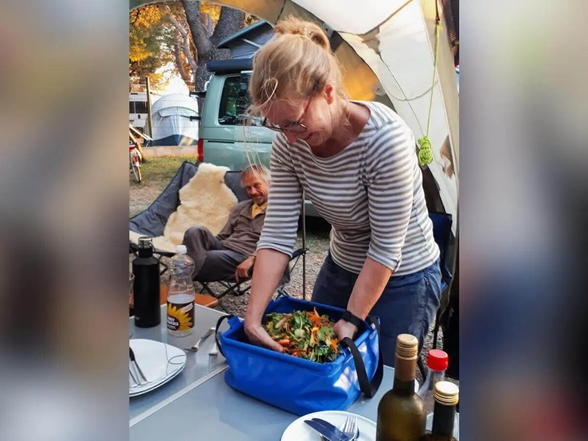 Eine Frau mischt Salat am Campingtisch; Geschirr, Weinflaschen und Zelte rahmen die Szene. | © DAV Markt Schwaben | Foto Martin und Silvia Mack