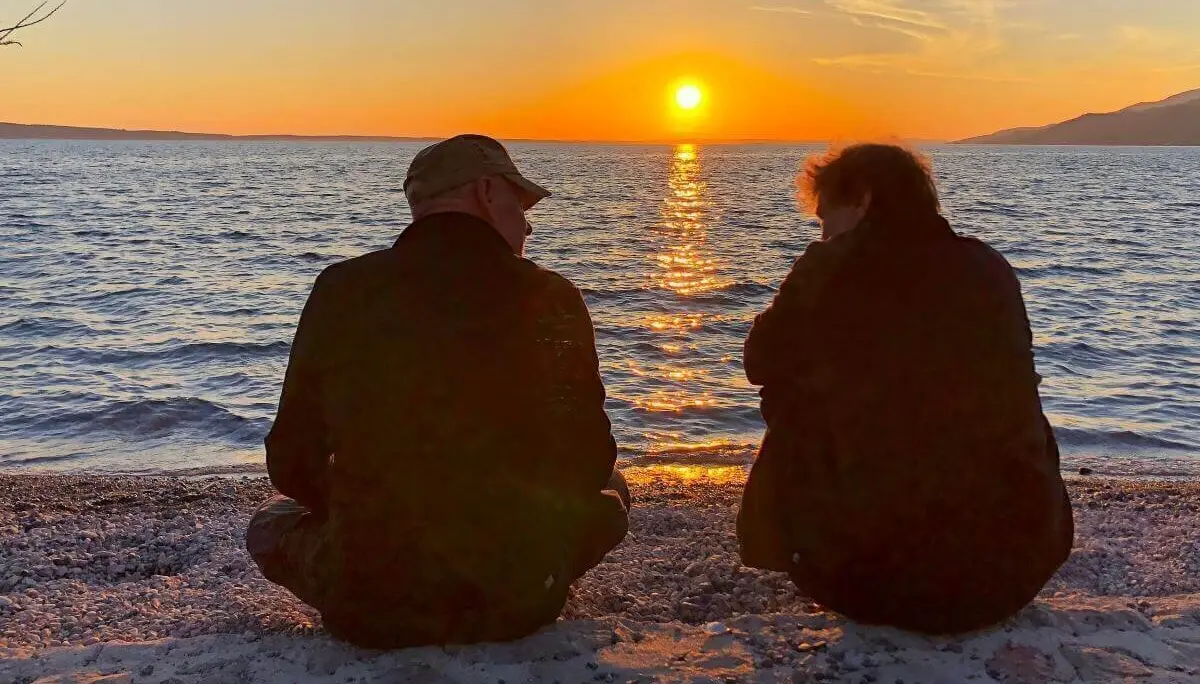 Zwei Personen sitzen am Kiesstrand und blicken auf den Sonnenuntergang über dem ruhigen Wasser. | © DAV Markt Schwaben | Foto Hildegard Petschik