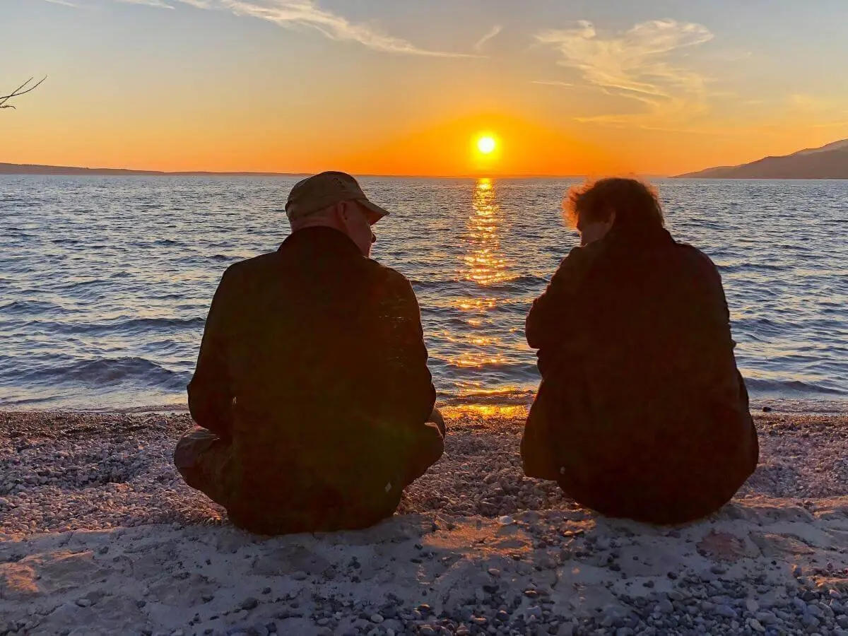 Zwei Personen sitzen am Kiesstrand und blicken auf den Sonnenuntergang über dem ruhigen Wasser. | © DAV Markt Schwaben | Foto Hildegard Petschik