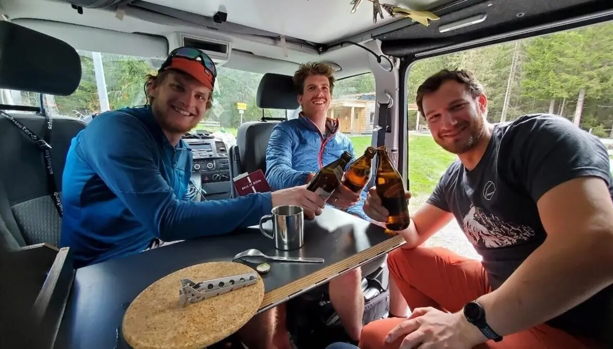 Drei Bergsteiger stoßen im Camper mit Bier auf ihre Tour an – entspannte Stimmung nach dem Abenteuer, Waldkulisse im Hintergrund. | © DAV Markt Schwaben | Foto: Walter Kressirer