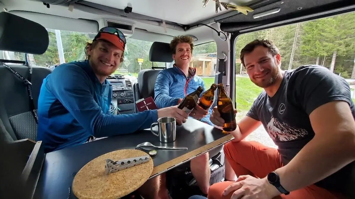 Drei Bergsteiger stoßen im Camper mit Bier auf ihre Tour an – entspannte Stimmung nach dem Abenteuer, Waldkulisse im Hintergrund. | © DAV Markt Schwaben | Foto: Walter Kressirer