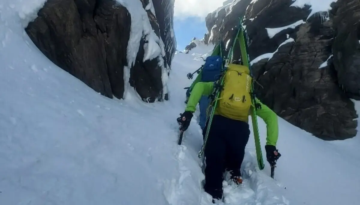 Ein Bergsteiger kämpft sich mit Ski am Rucksack durch tiefen Schnee in einer schmalen Ausstiegsrinne zwischen felsigen Flanken – Sonne und blauer Himmel im Hintergrund. | © DAV Markt Schwaben | Foto: Walter Kressirer
