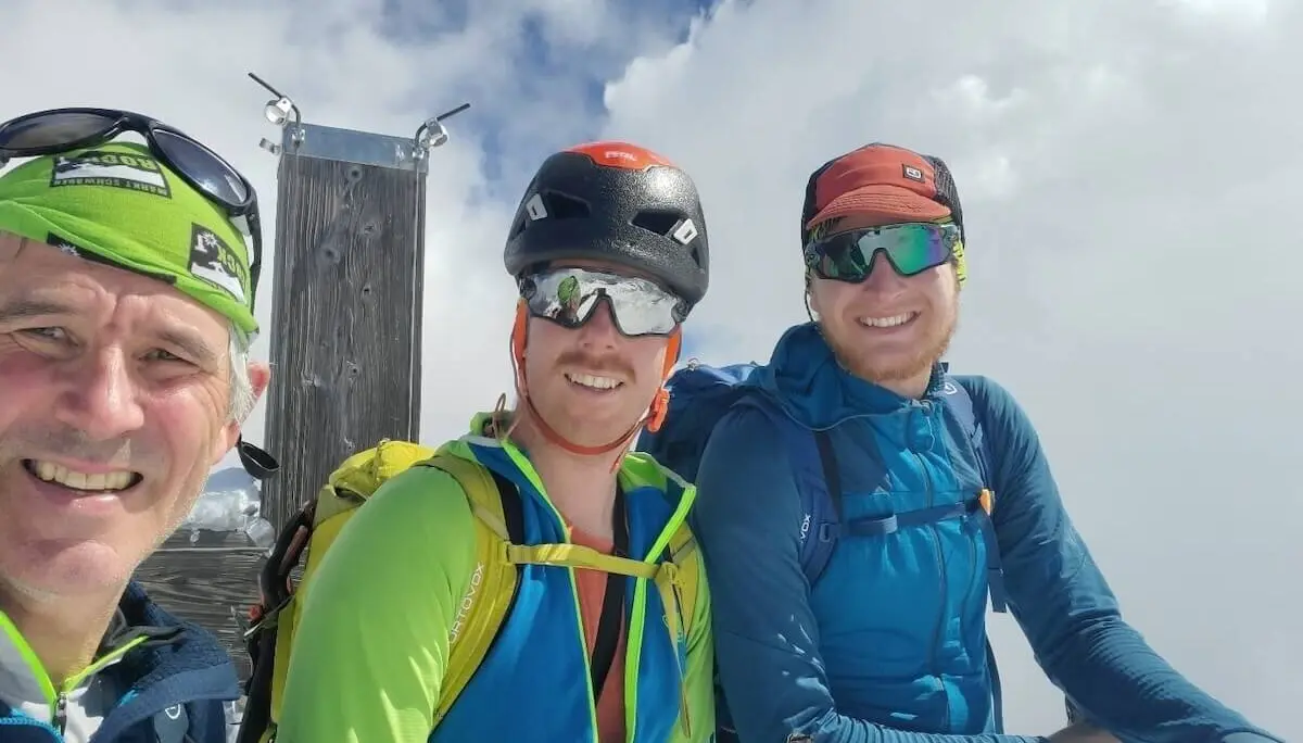 Drei Bergsteiger mit Helmen und Rucksäcken stehen oder sitzen am Gipfel des Schareck (3122 m) vor einer Holzkonstruktion – Wolkenmeer im Hintergrund, sichtlich stolz nach dem erfolgreichen Aufstieg. | © DAV Markt Schwaben | Foto: Walter Kressirer