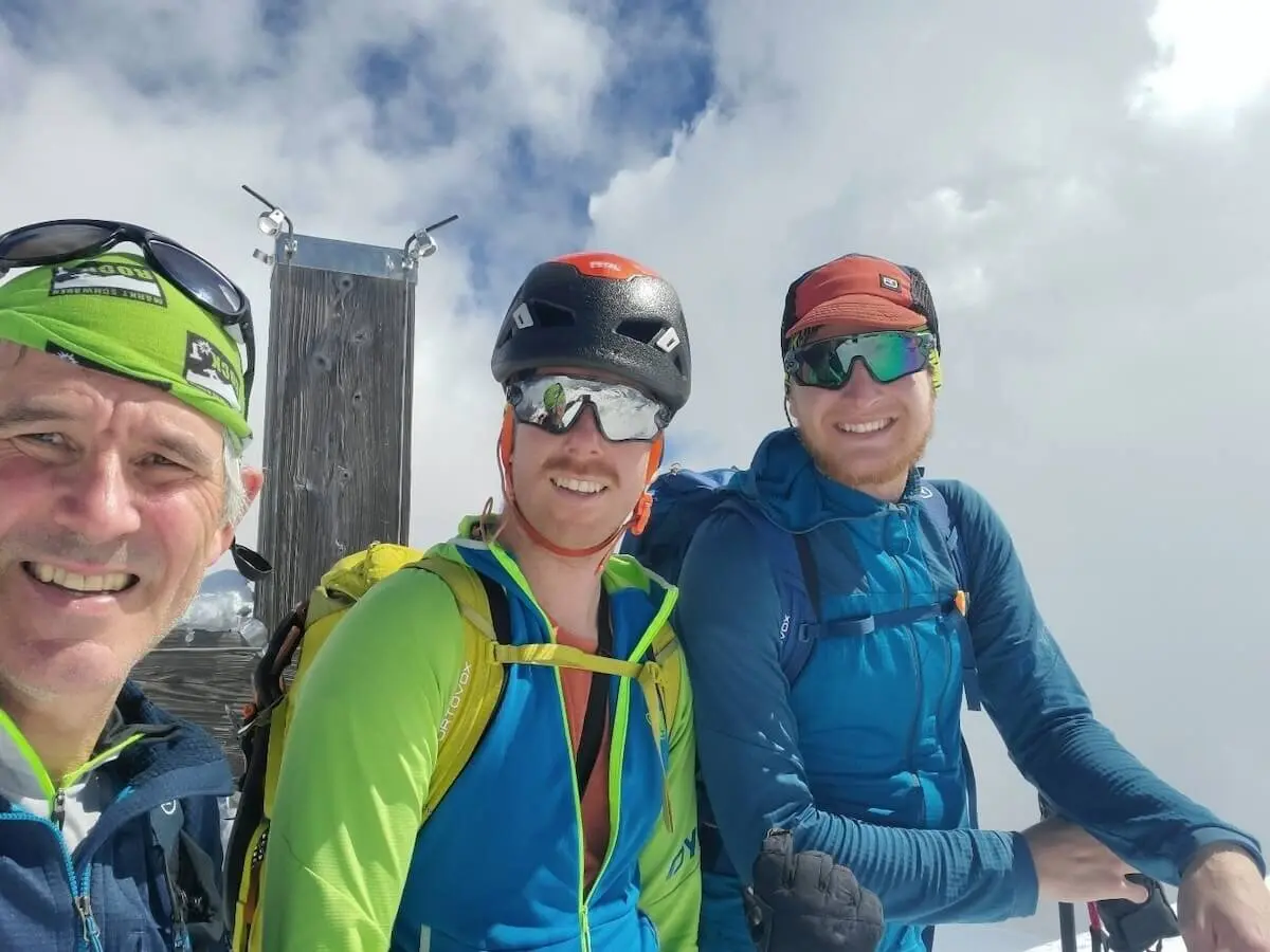 Drei Bergsteiger mit Helmen und Rucksäcken stehen oder sitzen am Gipfel des Schareck (3122 m) vor einer Holzkonstruktion – Wolkenmeer im Hintergrund, sichtlich stolz nach dem erfolgreichen Aufstieg. | © DAV Markt Schwaben | Foto: Walter Kressirer