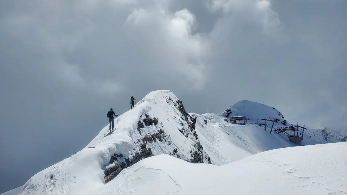 Zwei Bergsteiger folgen dem verschneiten Südgrat vom Gipfel des Schareck – im Hintergrund die Baumbachspitze und die Bergstation des Skigebiets, dahinter weite Firnhänge unter leicht bewölktem Himmel. | © DAV Markt Schwaben | Foto: Walter Kressirer