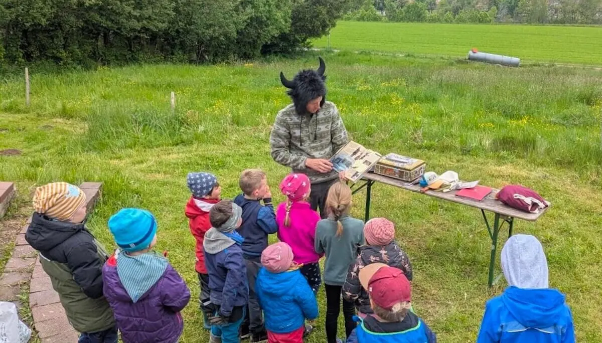 Eine Person mit einem Hut mit Hörnern zeigt einer Gruppe von Kindern draußen ein Buch. Sie stehen um einen Tisch mit Materialien. | © DAV Markt Schwaben | Foto: Familiengruppe