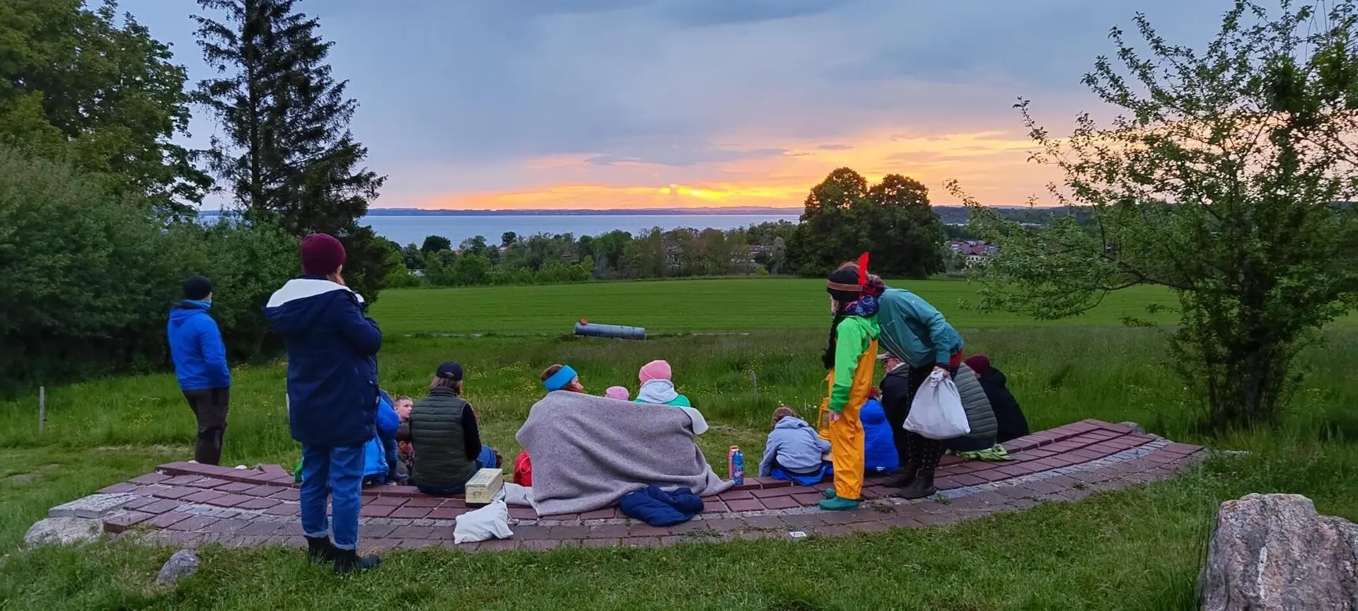 Eine Gruppe von Menschen sitzt auf halbrunden Steinstufen im Freien und posiert für ein Foto. Die Umgebung ist grün und von Bäumen umgeben. | © DAV Markt Schwaben | Foto: Familiengruppe