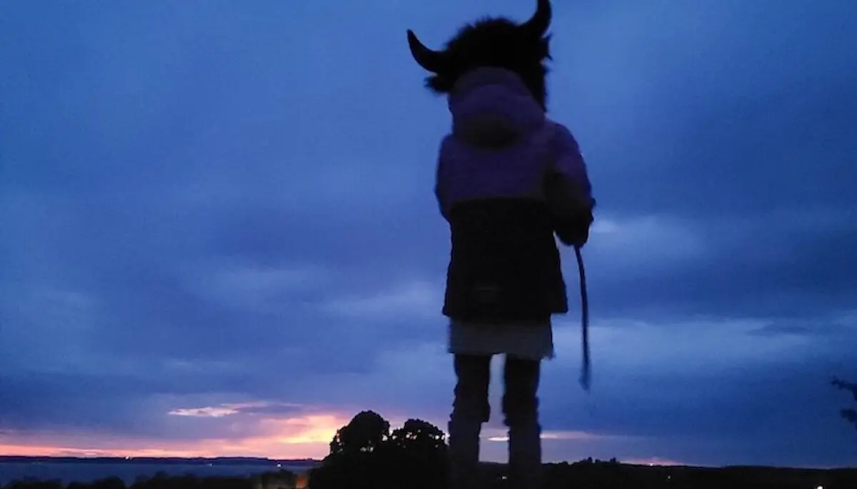 Eine Person mit einem Hut mit Hörnern steht bei Sonnenuntergang auf einem Hügel. Der Himmel leuchtet in warmen Blau- und Orangetönen. | © DAV Markt Schwaben | Foto: Familiengruppe