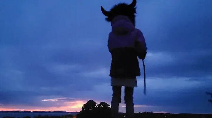 Eine Person mit einem Hut mit Hörnern steht bei Sonnenuntergang auf einem Hügel. Der Himmel leuchtet in warmen Blau- und Orangetönen. | © DAV Markt Schwaben | Foto: Familiengruppe