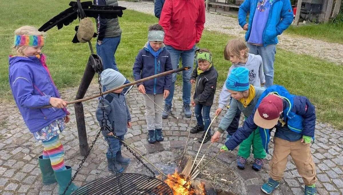 Eine Gruppe von Kindern sitzt oder steht um eine Feuerstelle im Freien. Sie tragen wetterfeste Kleidung und scheinen in das gemeinsame Erlebnis vertieft zu sein. | © DAV Markt Schwaben | Foto: Familiengruppe