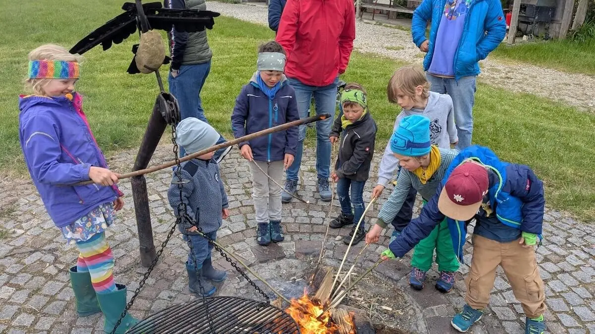 Eine Gruppe von Kindern sitzt oder steht um eine Feuerstelle im Freien. Sie tragen wetterfeste Kleidung und scheinen in das gemeinsame Erlebnis vertieft zu sein. | © DAV Markt Schwaben | Foto: Familiengruppe