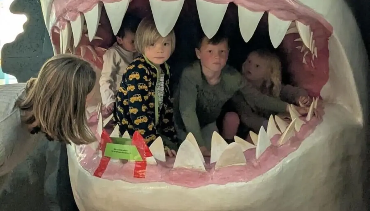 Drei Kinder sitzen im offenen Maul eines riesigen Haifischmodells, das als interaktives Museumsexponat gestaltet ist. | © DAV Markt Schwaben | Familiengruppe