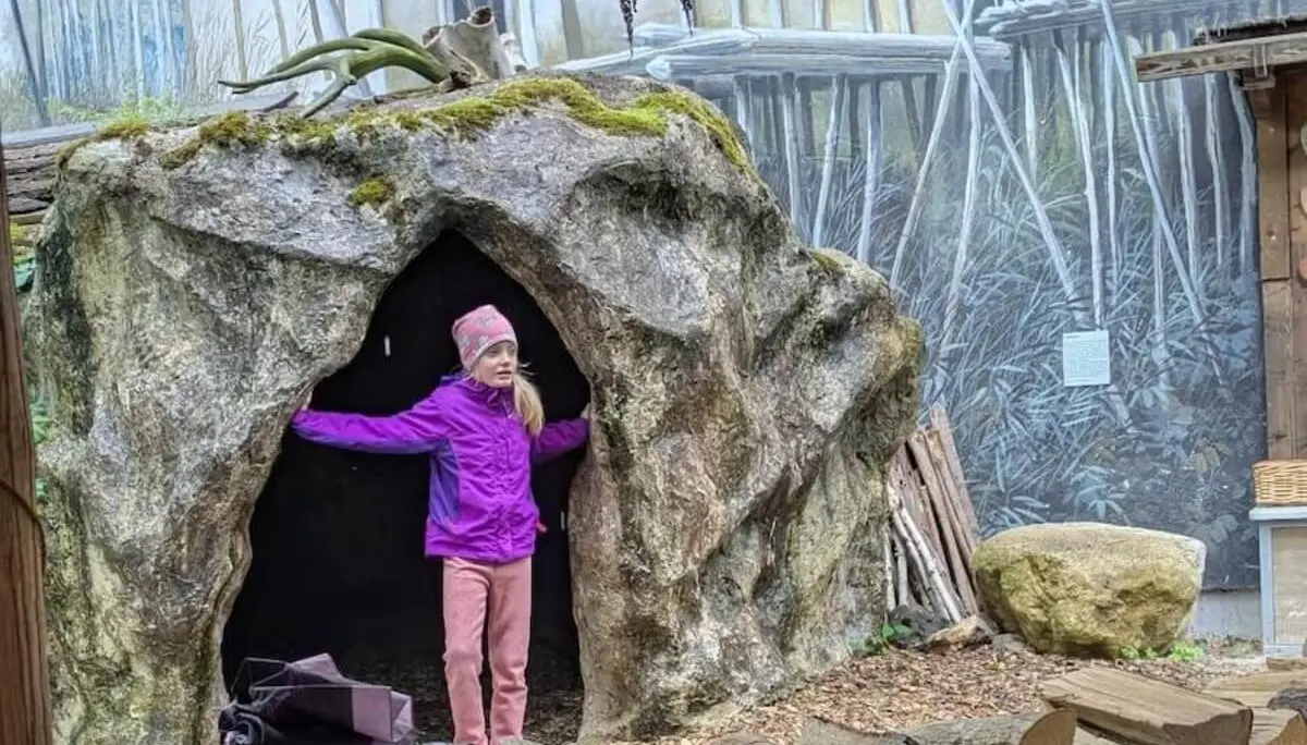 Ein Kind in lilafarbener Jacke und pinker Hose steht vor dem Eingang einer Höhlenstruktur aus Stein. Neben ihr liegt ein ordentlich gestapelter Holzstoß, während im Hintergrund ein Wandgemälde eine rustikale Szene mit Holzbauten und Vegetation zeigt. | © DAV Markt Schwaben | Familiengruppe