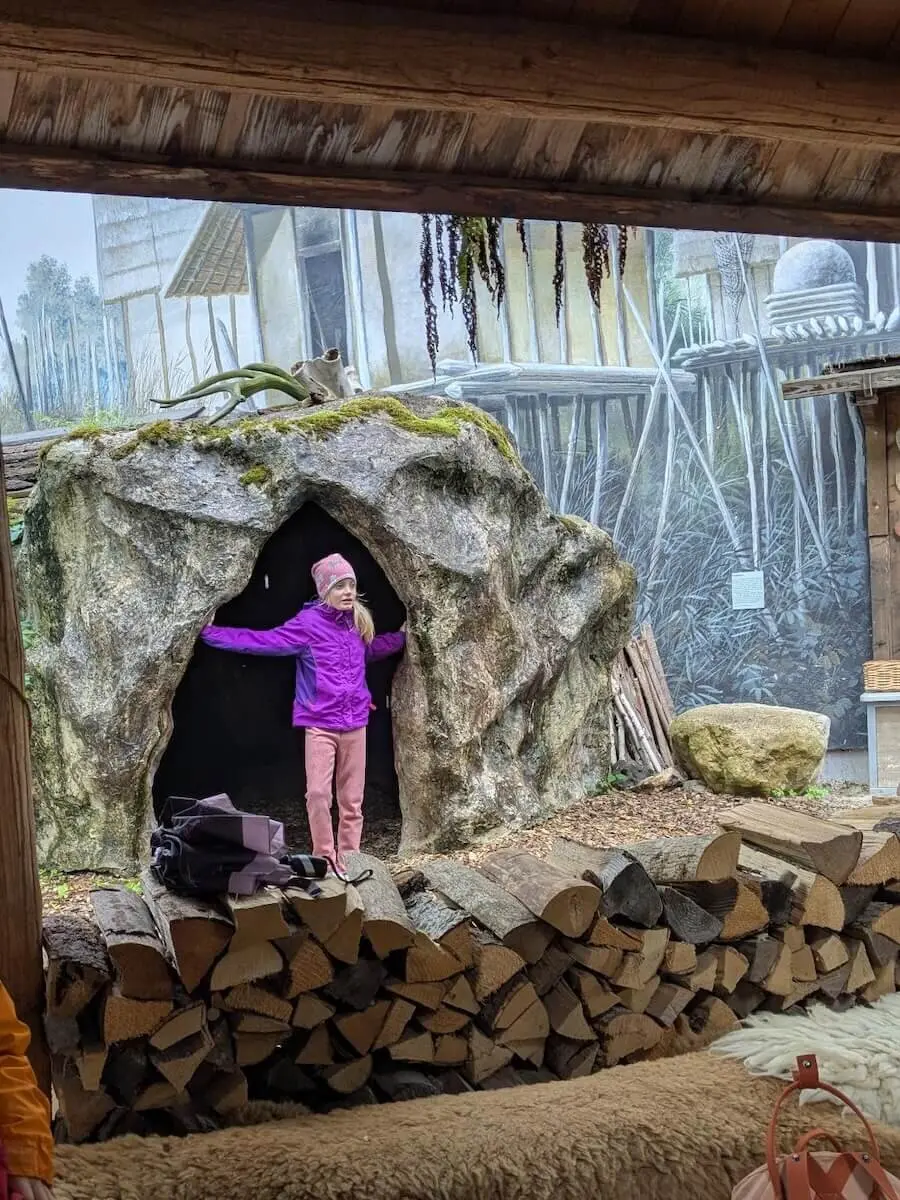 Ein Kind in lilafarbener Jacke und pinker Hose steht vor dem Eingang einer Höhlenstruktur aus Stein. Neben ihr liegt ein ordentlich gestapelter Holzstoß, während im Hintergrund ein Wandgemälde eine rustikale Szene mit Holzbauten und Vegetation zeigt. | © DAV Markt Schwaben | Familiengruppe