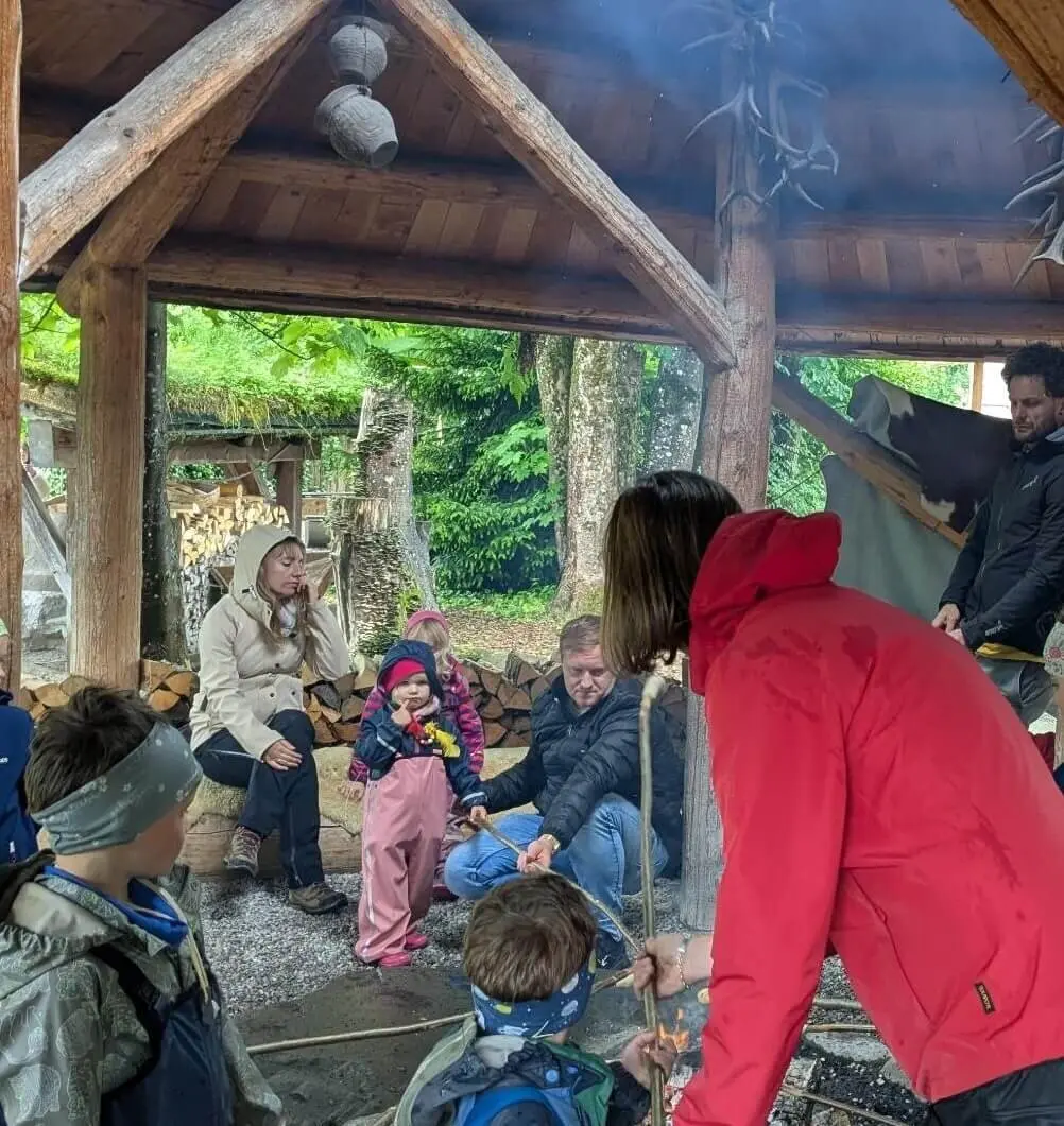 Eine Gruppe von Menschen, darunter Kinder und Erwachsene, versammelt sich unter einer hölzernen Überdachung im Wald. Einige halten Stöcke über ein Feuer, während im Hintergrund ein Stapel Brennholz zu sehen ist. Die Szene vermittelt eine gemütliche und gemeinschaftliche Atmosphäre bei einem Outdoor-Erlebnis. | © DAV Markt Schwaben | Familiengruppe