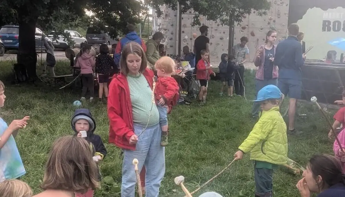 Kinder rösten Marshmallows am Lagerfeuer, im Hintergrund klettern andere an einer Wand – lebhaftes Outdoor-Erlebnis mit Spiel und Genuss. | © DAV Markt Schwaben | Foto Silke Titze