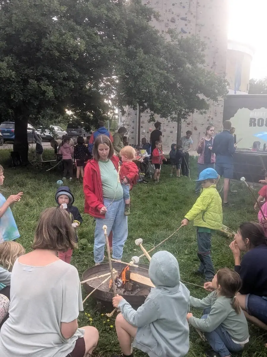 Kinder rösten Marshmallows am Lagerfeuer, im Hintergrund klettern andere an einer Wand – lebhaftes Outdoor-Erlebnis mit Spiel und Genuss. | © DAV Markt Schwaben | Foto Silke Titze