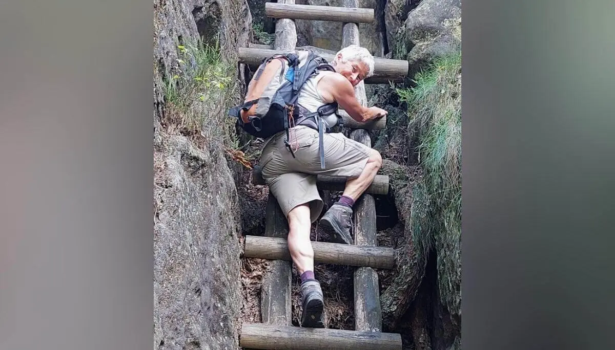 Person steigt senkrechte Holzleiter zwischen zwei Felswänden hinauf, umgeben von Moos und natürlichem Gelände. | © DAV Markt Schwaben | Foto Lutz Gründel