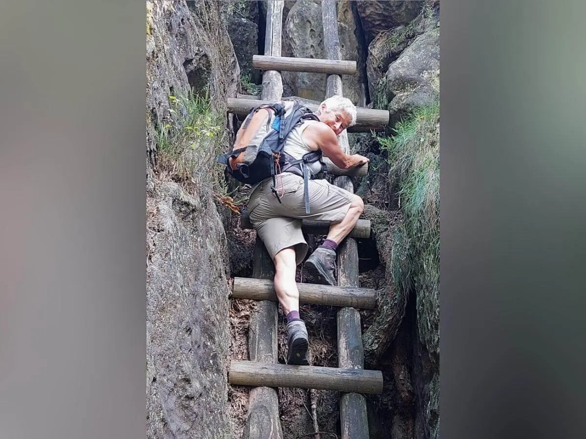 Person steigt senkrechte Holzleiter zwischen zwei Felswänden hinauf, umgeben von Moos und natürlichem Gelände. | © DAV Markt Schwaben | Foto Lutz Gründel
