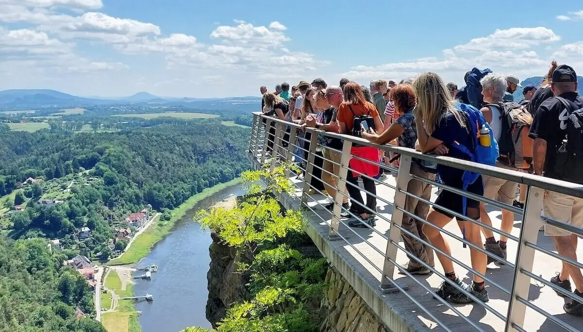 Gruppe auf Plattform über Felswand, Blick ins grüne Tal mit Fluss, Häusern und Hügeln unter blauem Himmel. | © DAV Markt Schwaben | Foto Michaela Haaß