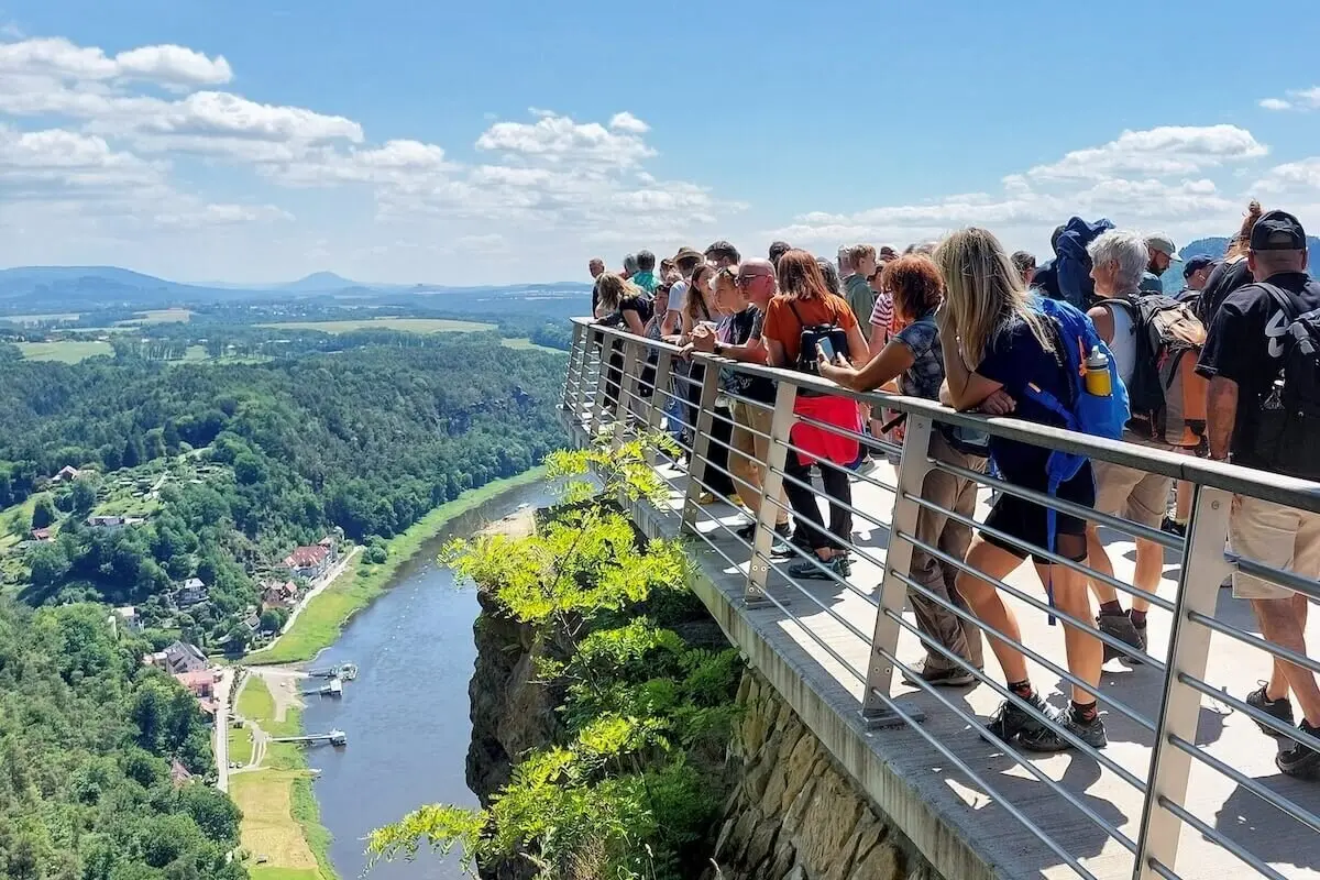 Gruppe auf Plattform über Felswand, Blick ins grüne Tal mit Fluss, Häusern und Hügeln unter blauem Himmel. | © DAV Markt Schwaben | Foto Michaela Haaß