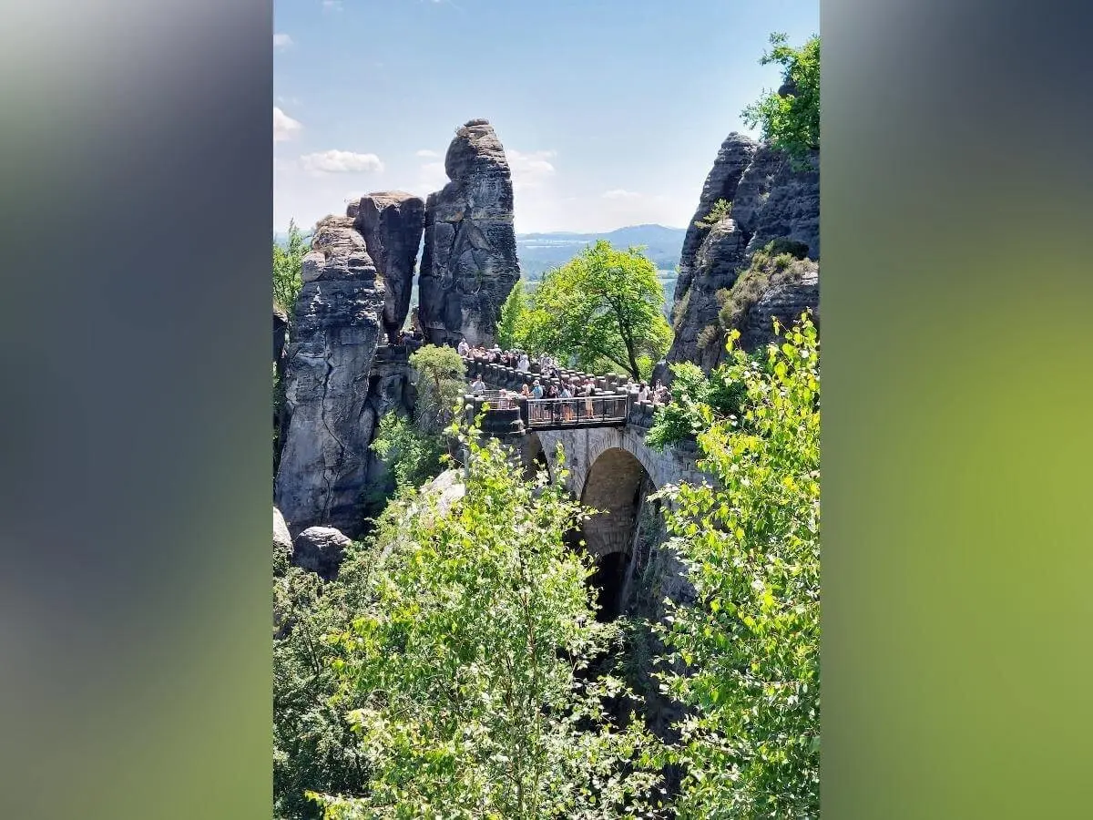 Zahlreiche Menschen überqueren steinerne Basteibrücke zwischen Felsen, umgeben von Wald und blauem Himmel. | © DAV Markt Schwaben | Foto Michaela Haaß