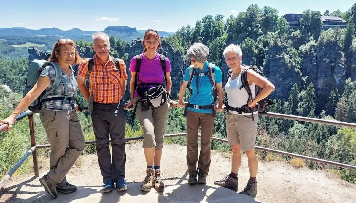 Fünf Wandernde stehen am Geländer, dahinter bewaldete Berge, Felsen und ein Gebäude auf dem Gipfel. | © DAV Markt Schwaben | Foto Michaela Haaß