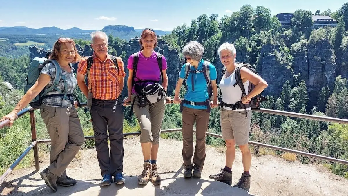 Fünf Wandernde stehen am Geländer, dahinter bewaldete Berge, Felsen und ein Gebäude auf dem Gipfel. | © DAV Markt Schwaben | Foto Michaela Haaß
