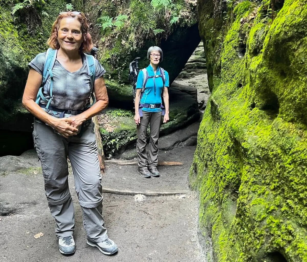Zwei Personen stehen auf Steinpfad zwischen moosbedeckten Felsen, umgeben von Farnen und dichter Vegetation. | © DAV Markt Schwaben | Foto Edith Zerrle