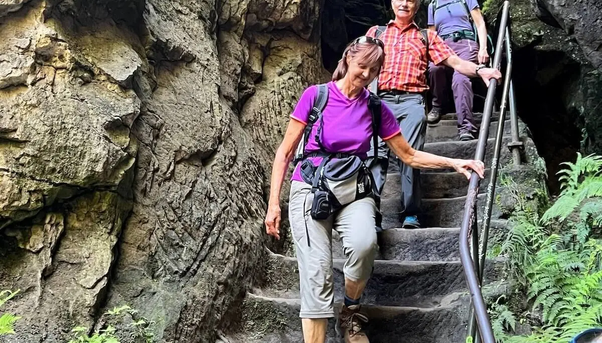 Drei Personen steigen schmale Steintreppe mit Geländer hinab, umgeben von rauem Fels und etwas Grün. | © DAV Markt Schwaben | Foto Edith Zerrle