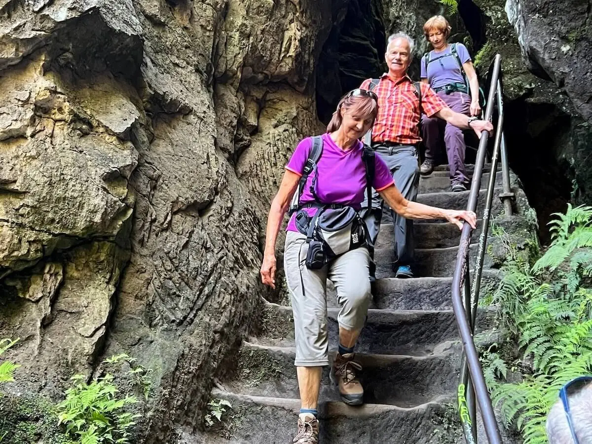 Drei Personen steigen schmale Steintreppe mit Geländer hinab, umgeben von rauem Fels und etwas Grün. | © DAV Markt Schwaben | Foto Edith Zerrle