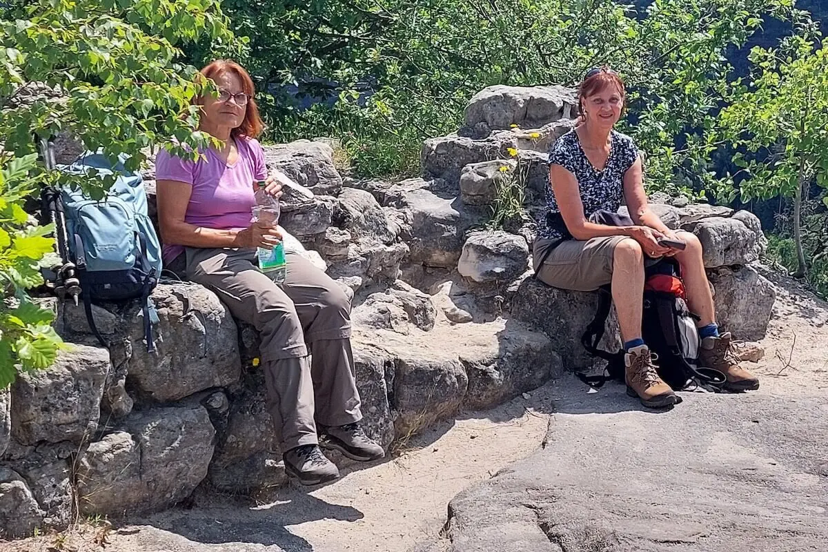 Zwei Personen sitzen auf Felsen mit gelben Blumen, umgeben von Grün, beide tragen Wanderausrüstung. | © DAV Markt Schwaben | Foto Lutz Gründel