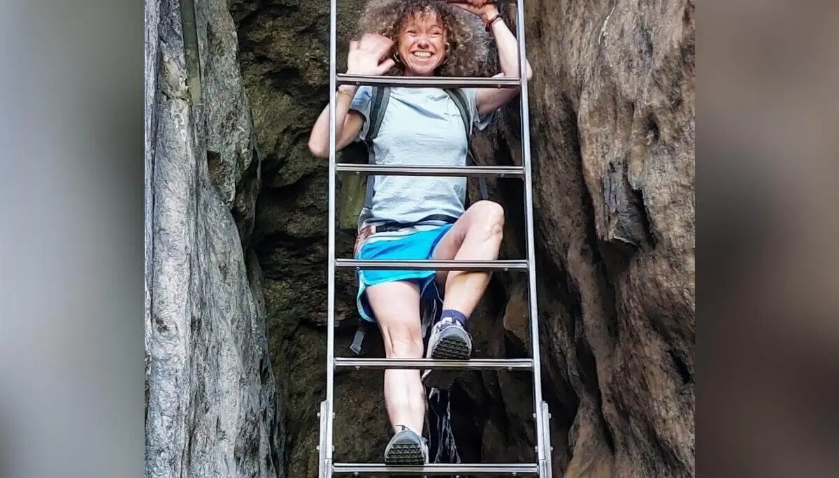 Person winkt beim Klettern auf Metallleiter zwischen Felsen, lächelt, umgeben von steilem Gelände. | © DAV Markt Schwaben | Foto Lutz Gründel