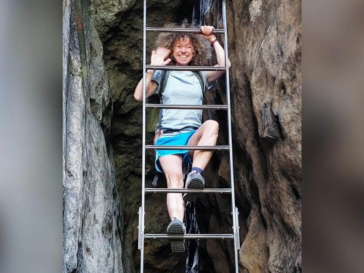 Person winkt beim Klettern auf Metallleiter zwischen Felsen, lächelt, umgeben von steilem Gelände. | © DAV Markt Schwaben | Foto Lutz Gründel