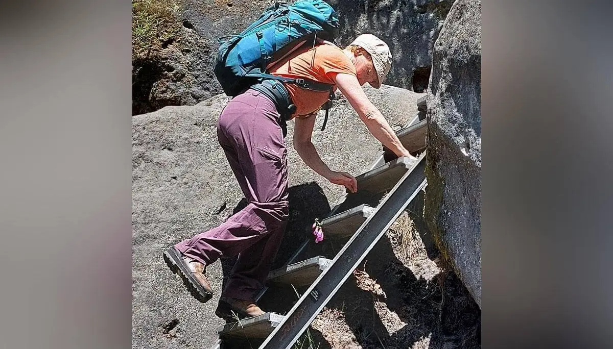 Person mit orangefarbenem Shirt und Rucksack erklimmt Metallleiter zwischen steilen Felsen. | © DAV Markt Schwaben | Foto Lutz Gründel